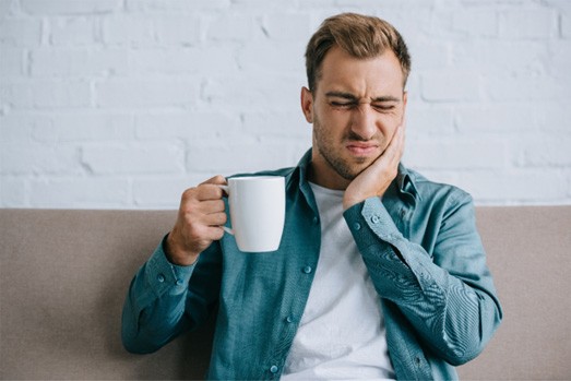 Man on couch squinting in pain touching jaw holding white mug