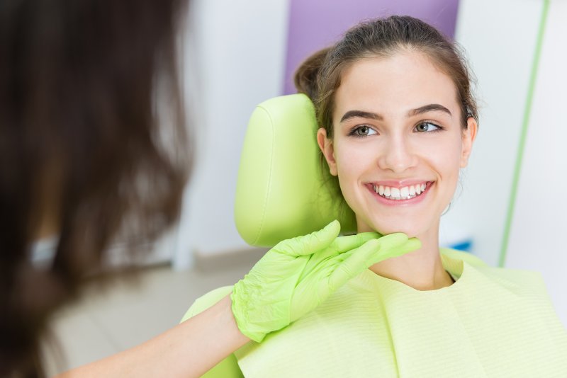 Young woman smiling in dental chair as dentist holds her chin with gloved hand